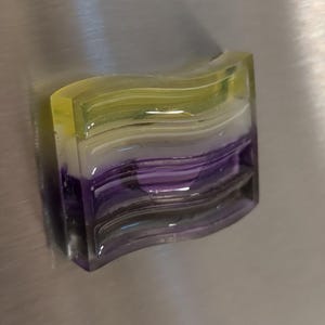 May include: A multi-layered, wavy magnet in shades of yellow, white, and purple. The magnet, made of a clear, glossy material, is attached to a metallic surface. The design is abstract and contemporary.