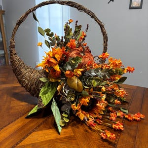 May include: A woven brown twig cornucopia basket filled with an arrangement of artificial orange and yellow flowers, green eucalyptus leaves, and a textured orange pumpkin. The decorative piece features a large circular handle made of twisted vines.