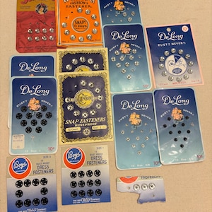 May include: A collection of vintage sewing snap fasteners on original cardboard display cards. Brands include DeLong, Wilsnap, Starsnap, Clinton, and Boye. The cards feature various counts of silver and black metal snaps, with printed text detailing brand names, rustproof features, and sizes.