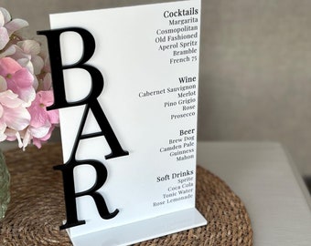 May include: A white sign with black lettering that says "BAR" in large letters. The sign lists a variety of cocktails, wines, beers, and soft drinks.