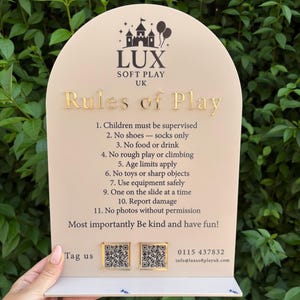 May include: A beige sign with gold lettering and the LUX SOFT PLAY UK logo. The sign details rules of play, such as supervision, no shoes, and no rough play. Two QR codes and contact details are at the bottom.