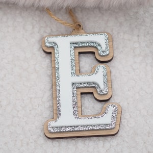 May include: Wooden letter "E" decoration with a white centre and silver glitter trim. The letter is layered on a wooden base and has a natural twine string for hanging. The ornament is set against a soft, textured background.