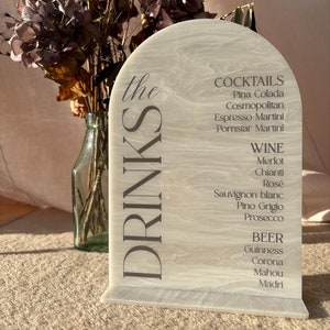 May include: A white arch-shaped sign with the text "the DRINKS" in black lettering. The sign lists a variety of cocktails, wines, and beers, including Pina Colada, Merlot, and Guinness.