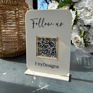 May include: A white acrylic sign with the words "Follow us" in black script and a QR code in a gold frame. The Etsy shop name "EttyDesigns" is printed at the bottom. The sign is displayed on a blue surface.