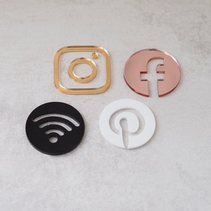 May include: Four social media icons made of acrylic. The icons are in the shape of a camera, a letter f, a wifi symbol, and a letter p. The camera icon is gold, the letter f is rose gold, the wifi symbol is black, and the letter p is white.