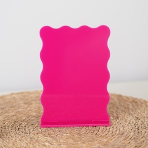 May include: A bright pink acrylic sign with a wavy edge. The sign is standing on a woven mat.