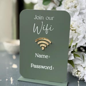 Op de afbeelding: Een groen rechthoekig bord met afgeronde hoeken, met de woorden "Join our Wifi" in witte letters. Daaronder een goudkleurig wifi-symbool. Het bord bevat velden voor "Name:" en "Password:" in witte tekst.