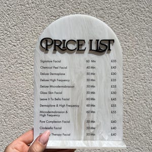 Puede incluir: Una lista de precios de acr&iacute;lico blanco y negro veteado con una parte superior curvada. El texto negro dice "PRICE LIST" encima de una lista de tratamientos faciales, incluyendo Signature Facial y Chemical Peel Facial.