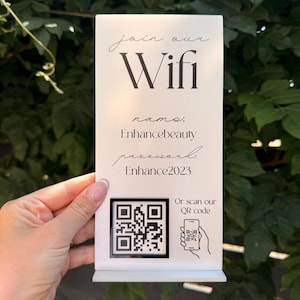 May include: A rectangular white sign with black text and a QR code. The text reads: join our Wifi, name: Enhancebeauty, password: Enhance2023, and Or scan our QR code with a small icon of a hand holding a phone. The sign stands on a flat base.