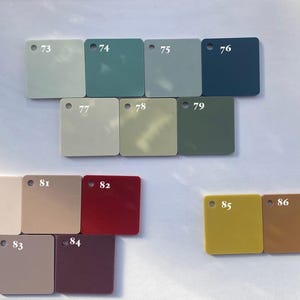 Op de afbeelding: Een verzameling vierkante kleurstalen in verschillende tinten, waaronder lichtgroen, blauwgroen, grijs en diepblauw. Andere kleuren zijn beige, olijfgroen, rood en mosterdgeel. Elke staal is genummerd van 73 tot 86.