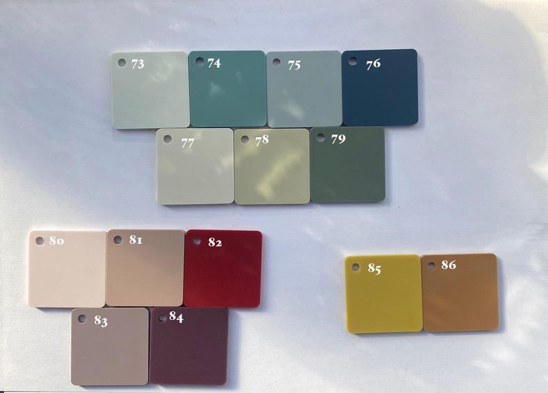 Op de afbeelding: Een verzameling vierkante kleurstalen in verschillende tinten, waaronder lichtgroen, blauwgroen, grijs en diepblauw. Andere kleuren zijn beige, olijfgroen, rood en mosterdgeel. Elke staal is genummerd van 73 tot 86.