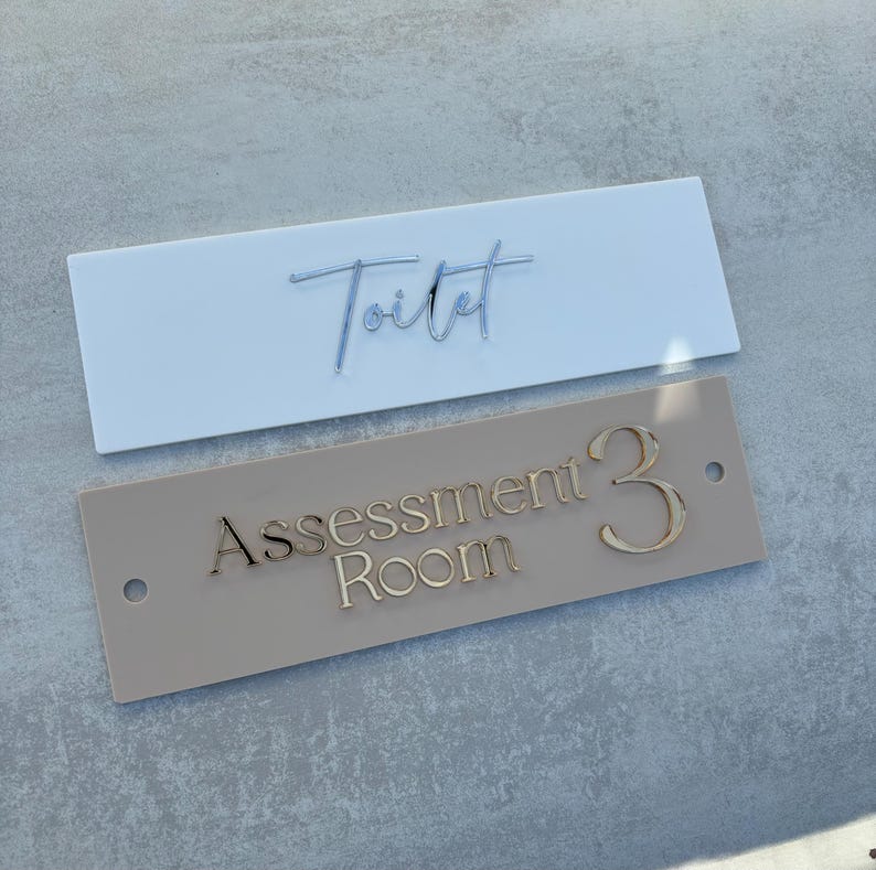 May include: Two rectangular signs with gold lettering. The white sign says "Toilet" in cursive. The beige sign says "Assessment Room 3".