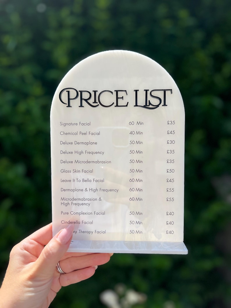 Puede incluir: Una lista de precios de acr&iacute;lico blanco con una parte superior curvada, con texto negro que dice "PRICE LIST". La lista detalla varios tratamientos faciales, incluyendo Signature Facial, Chemical Peel Facial y Deluxe Dermaplane, con los tiempos asociados.