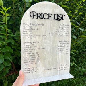 Puede incluir: Una lista de precios blanca y crema con texto negro, que enumera los servicios de peluquer&iacute;a y los precios. El letrero lo sostiene una persona. El texto incluye servicios como cortes de pelo, coloraci&oacute;n y extensiones de cabello.