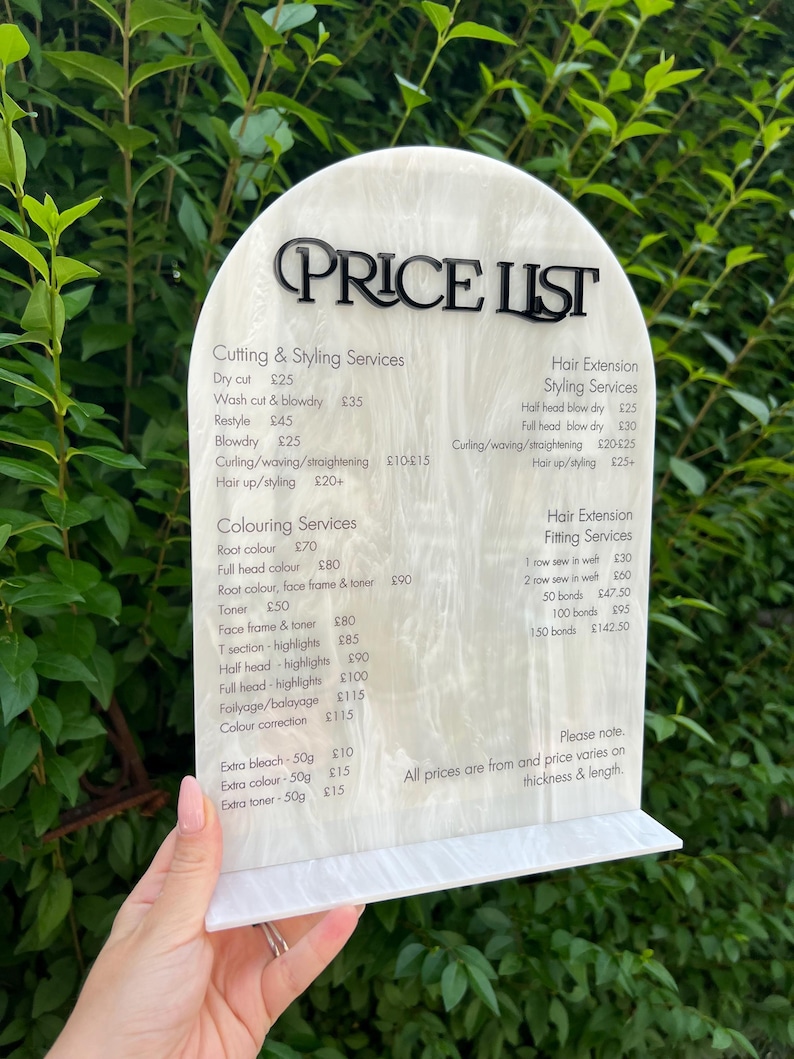 Puede incluir: Una lista de precios blanca y crema con texto negro, que enumera los servicios de peluquer&iacute;a y los precios. El letrero lo sostiene una persona. El texto incluye servicios como cortes de pelo, coloraci&oacute;n y extensiones de cabello.