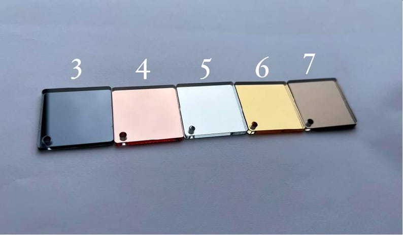 Op de afbeelding: Zes vierkante, spiegelende acrylplaten in verschillende kleuren: donkergrijs, ros&eacute;goud, zilver, goud en brons. Elk stuk heeft een klein gaatje en is genummerd van 3 tot 7.