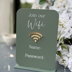 Op de afbeelding: Een saliegroen WiFi-bord met een goudkleurig WiFi-symbool. Op het bord staat "Join our Wifi" en er zijn ruimtes voor "Name:" en "Password:" in witte tekst. Het bord staat op een bijpassende basis.