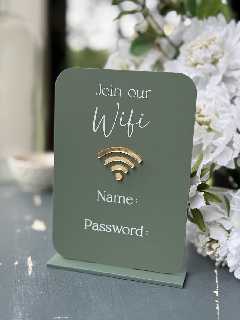 Op de afbeelding: Een saliegroen WiFi-bord met een goudkleurig WiFi-symbool. Op het bord staat "Join our Wifi" en er zijn ruimtes voor "Name:" en "Password:" in witte tekst. Het bord staat op een bijpassende basis.
