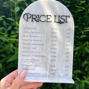 Puede incluir: Una lista de precios blanca y gris con efecto m&aacute;rmol y texto negro. El letrero enumera varios tratamientos faciales, incluyendo Signature Facial, Chemical Peel Facial y Deluxe Dermaplane. El letrero se sostiene frente a un fondo de hojas verdes.