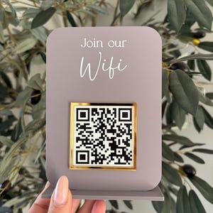 May include: A mauve-coloured WiFi sign with the words "Join our Wifi" in white script. A gold-framed QR code is centered on the sign. The sign is displayed against a backdrop of olive branches with green leaves and black olives.