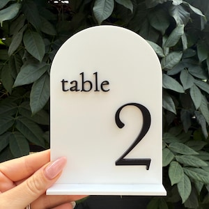 May include: A white table number with a black number 2 and the word "table" in black. The table number has an arched top and a small base, perfect for weddings or events.