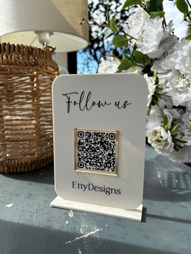 May include: A white acrylic sign with the words "Follow us" in black script and a QR code in a gold frame. The Etsy shop name "EttyDesigns" is printed at the bottom. The sign is displayed on a blue surface.
