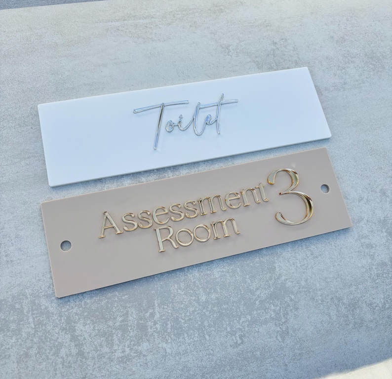 May include: Two rectangular signs, one white with the word "Toilet" in cursive script and one beige with the text "Assessment Room 3" in gold lettering.