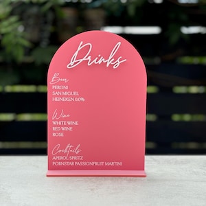 May include: Pink arch-shaped sign with white text listing drinks available: Beer, Peroni, San Miguel, Heineken 0.0%, Wine, White Wine, Red Wine, Rose, Cocktails, Aperol Spritz, Pornstar Passionfruit Martini.