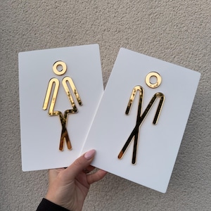 May include: Two white rectangular restroom signs with gold-colored stick figure symbols. One sign features a female figure, the other a male figure. The signs are held against a neutral-colored wall.