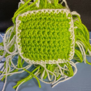 May include: A green crocheted square with a white border. The square has loose white and green yarn strands hanging from the edges.