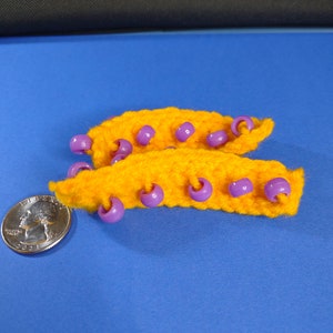 May include: Two orange crocheted strips with purple beads attached. The strips are about the size of a US quarter.