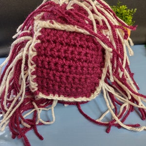 May include: A crocheted hat with a burgundy body and white trim. The hat has long white and burgundy fringe.