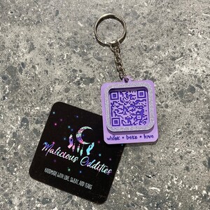 May include: A lilac QR code keyring with a silver glitter border and a silver chain. The keyring has the words "whisk • bake • love" printed on it. A black business card with the words "Malicious Oddities" is also in the image.