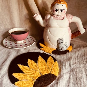 May include: Two brown and yellow sunflower-shaped pot holders with a white ceramic teapot shaped like a woman with a cat on her side. The teapot is on a white surface.