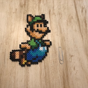 May include: A green and brown pixel art character with a green hat and a brown tail. The character is wearing blue overalls and is flying through the air.