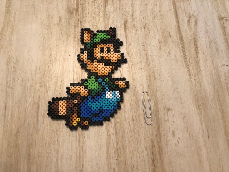 May include: A green and brown pixel art character with a green hat and a brown tail. The character is wearing blue overalls and is flying through the air.
