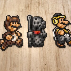 May include: Three pixel art figures made of colorful beads. The figures are of a raccoon Mario, a black knight, and a yellow-haired Mario.