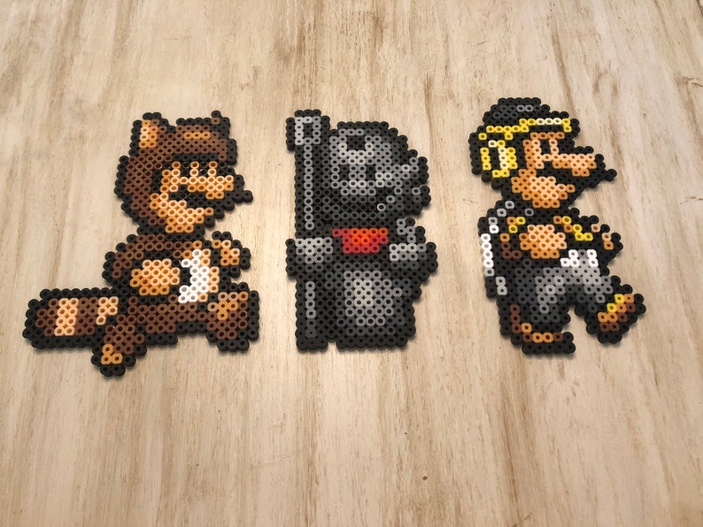 May include: Three pixel art figures made of colorful beads. The figures are of a raccoon Mario, a black knight, and a yellow-haired Mario.