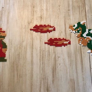 May include: A red and green pixel art figure of Mario, a video game character, stands on a wooden surface. Two red and orange pixel art fireballs are in the background, and a green and brown pixel art figure of Bowser, a video game character, is on the right side of the image.