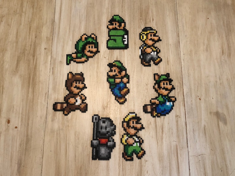 May include: Eight pixel art characters from the Super Mario Bros. franchise, including Mario, Luigi, Toad, and a raccoon Mario, are displayed on a wooden surface. The characters are made of colorful beads.