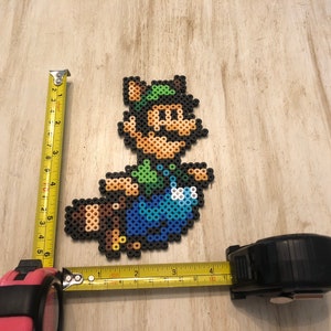May include: A pixel art character made of colorful beads, depicting a green-clad figure with a brown tail, a green hat, and a mustache. The figure is approximately 6 inches tall and 4 inches wide.