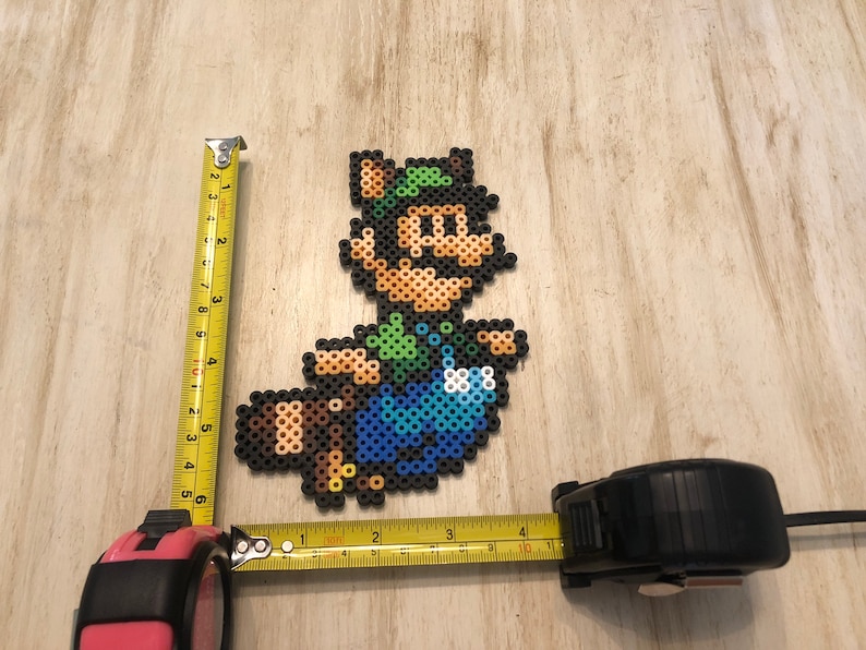 May include: A pixel art character made of colorful beads, depicting a green-clad figure with a brown tail, a green hat, and a mustache. The figure is approximately 6 inches tall and 4 inches wide.