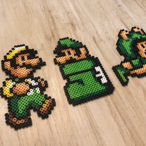 May include: Three pixel art figures made of green and brown perler beads. The figures are of a green and brown character with a yellow hat, a green character with a green hat, and a green character with a green hat and a brown face.