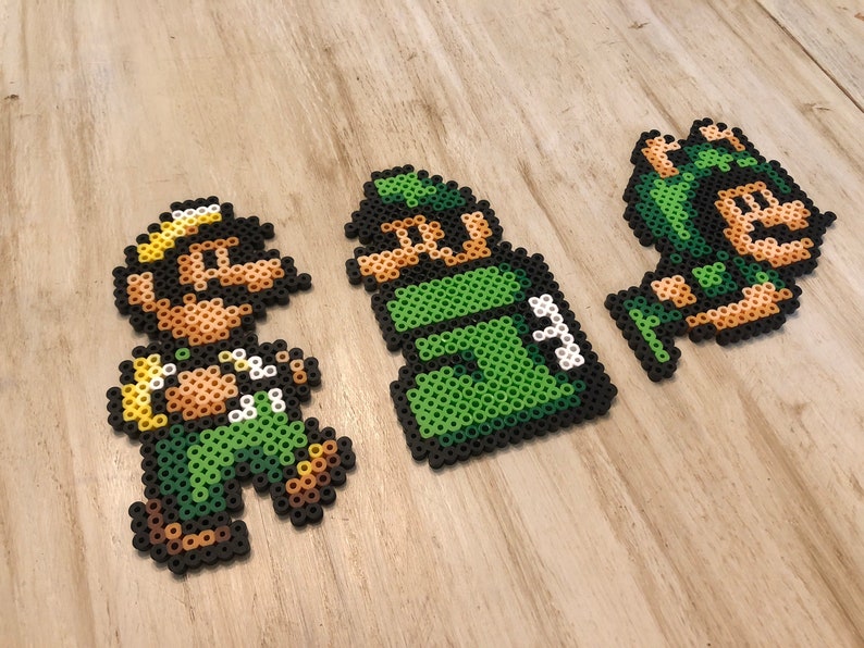 May include: Three pixel art figures made of green and brown perler beads. The figures are of a green and brown character with a yellow hat, a green character with a green hat, and a green character with a green hat and a brown face.