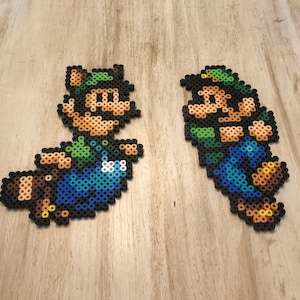 May include: Two pixel art figures of Mario characters, one is a brown raccoon Mario with a green hat and the other is a green and blue Luigi with a green hat. Both figures are made of colorful plastic beads.