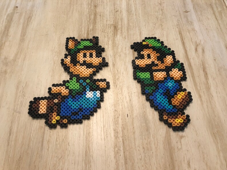 May include: Two pixel art figures of Mario characters, one is a brown raccoon Mario with a green hat and the other is a green and blue Luigi with a green hat. Both figures are made of colorful plastic beads.