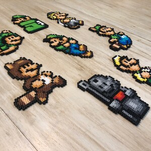 May include: A collection of eight pixel art characters made with colorful beads. The characters include a brown bear, a knight, a green and blue character, and several characters wearing green and blue clothing.