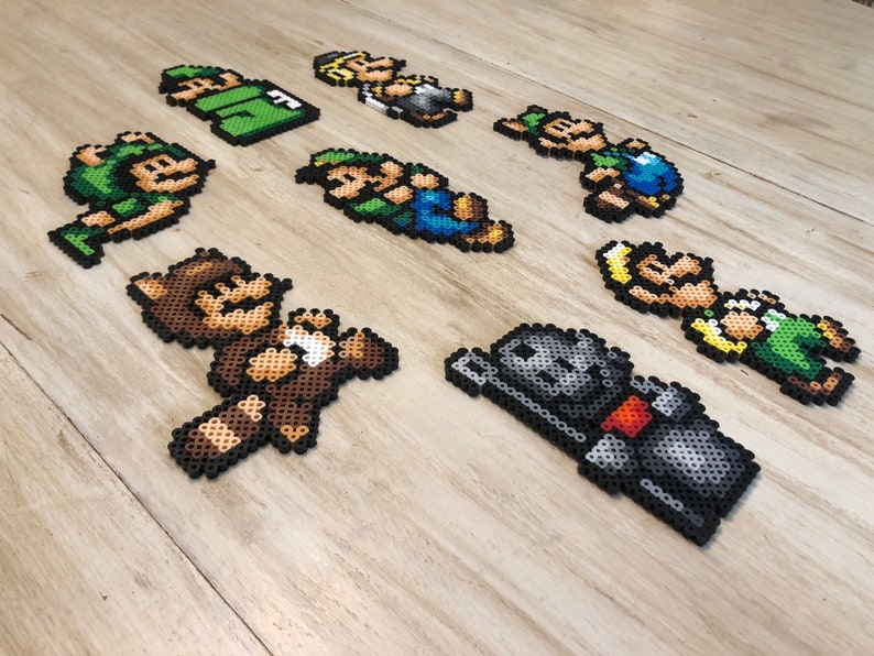 May include: A collection of eight pixel art characters made with colorful beads. The characters include a brown bear, a knight, a green and blue character, and several characters wearing green and blue clothing.
