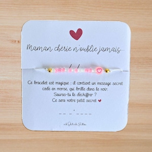 Peut inclure: Une carte blanche avec des cœurs roses et le texte "Maman chérie n'oublie jamais..." La carte est attachée à un bracelet en cordon blanc avec des perles roses et blanches et un charm en forme de cœur rose. Le texte sur la carte se lit comme suit : "Ce bracelet est magique : il contient un message secret codé en morse, qui brille dans le noir. Sauras-tu le déchiffrer ? Ce sera notre petit secret"