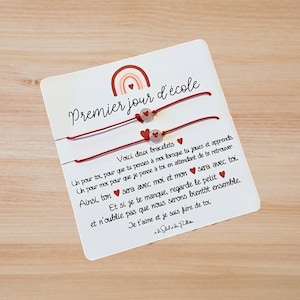 Peut inclure: Une carte avec le texte "Premier jour d'école" et deux bracelets en fil rouge. Chaque bracelet a une perle en forme de cœur et un petit charme en forme de cœur. Un arc-en-ciel est en haut de la carte.